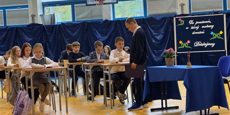 Powiększ grafikę: Uczniowie klas 5a i 5d w skupieniu uczestniczą w części artystycznej – jeden z uczniów trzyma mikrofon.