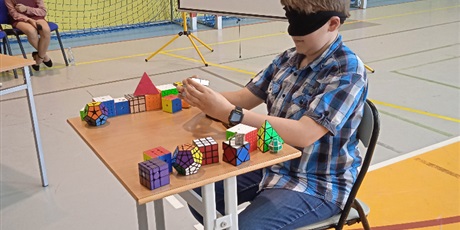 Powiększ grafikę: Uczeń z 5f siedzi przy stoliku i z czarną przepaską na oczach układa kostkę Rubika
