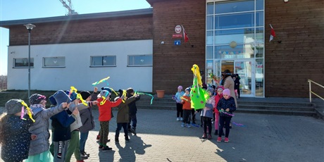 Powiększ grafikę: Dzieci ustawione w dwóch szeregach trzymają kolorowe chorągiewki i witają Panią Wiosnę. 